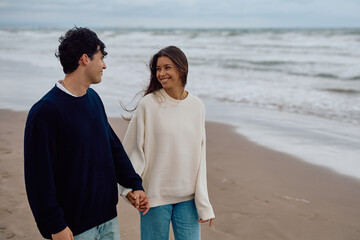 Young couple wearing cozy sweaters, holding hands and strolling along the beach on a cloudy day, enjoying their time together by the ocean