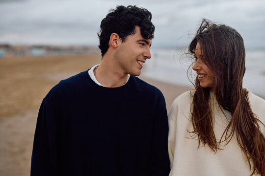 Young couple walking hand in hand along the beach, sharing smiles and gazing into each other's eyes, radiating love and happiness - Powered by Adobe