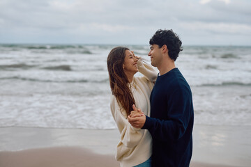 Young couple wearing cozy sweaters, holding hands and laughing joyfully together on the beach,...