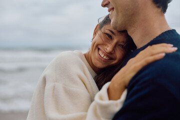 Fototapeta premium Young woman resting her head on her boyfriend's shoulder, smiling and hugging him on a beach with the sea in the background