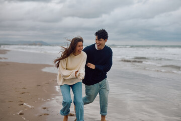 Happy couple running barefoot along the beach, laughing joyfully and enjoying carefree moments together under a cloudy autumn sky