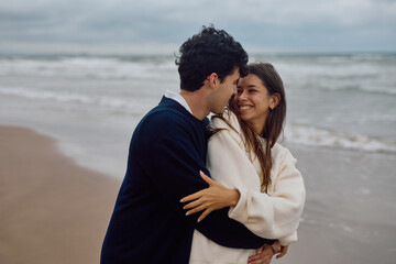 Fototapeta premium Romantic couple hugging and smiling on the beach with the ocean in the background during a cloudy day