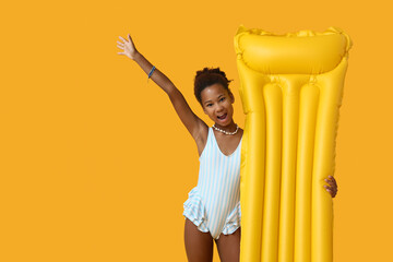 Cute little African-American girl in swimsuit with inflatable mattress on yellow background