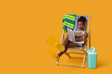 Cute little African-American girl in swimsuit and flippers reading magazine on deckchair against yellow background © Pixel-Shot