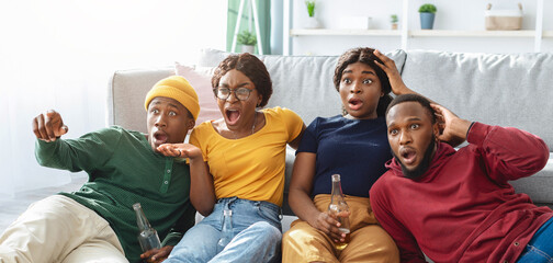 Shocked african american friends sitting on floor, watching horror movie and drinking beer at home, copy space. Astonished group of young black men and women spending time together, having party