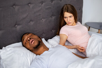 Millennial black man snoring and bothering his irritated sleepless wife in bed at home. Young Caucasian lady having trouble falling asleep, looking at her husband in disgust, cannot bear loud noise