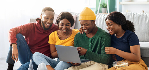 Happy black friends young men and women using laptop, sitting on floor in living room, drinking...