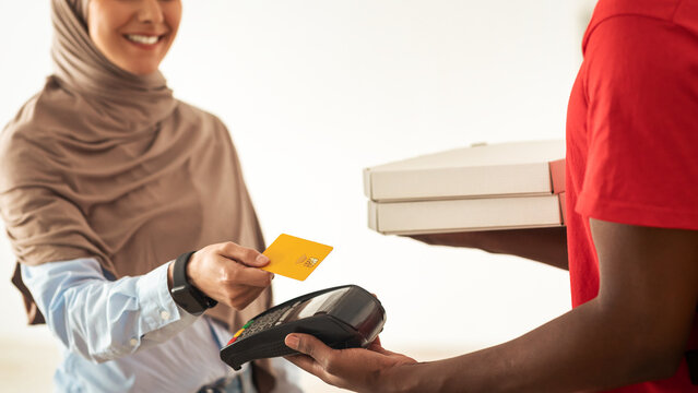 Delivery And Technology. Smiling muslim woman in hijab using credit card for contactless payment, black male courier in red uniform holding Point Of Sale atm machine in hand and pizza boxes