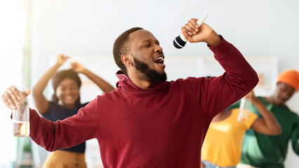 Funny black guy singing karaoke at home, holding bottle of beer, dancing and cheering group of...