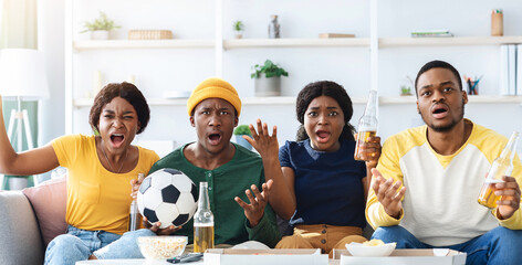 Furious african american friends drinking beer and eating snacks, watching football game on TV at...