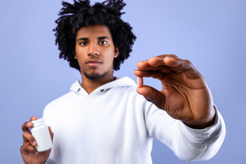 Black teenager holding jar of tablets and showing one pill over blue studio background, selective...