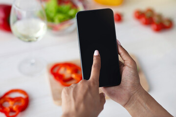 Social networks at home, mobile app and cooking with gadget. Millennial african american female use smartphone with blank screen in kitchen, at table with bright vegetables background, pov, mockup