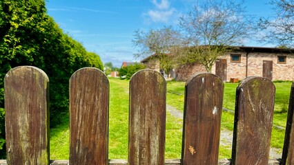 weathered wooden fence frames a view of a green lawn and a brick building under a bright blue sky with scattered clouds.