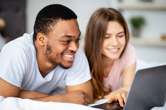 Affectionate multiracial couple spending time together at home, using laptop on bed. Happy black man and his Caucasian wife watching video, browsing web or having online conference