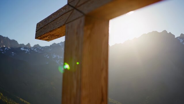 A wooden cross stands in front of misty mountain peaks with sunlight streaming through. The foreground features lens flare and the background has snow patches on the mountains. - Powered by Adobe