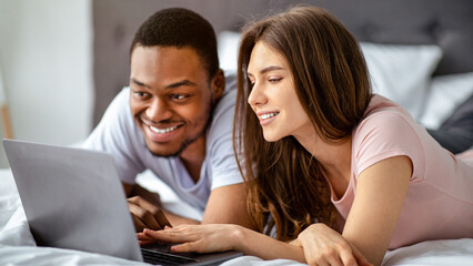 Interracial loving couple spending weekend together at home, using laptop while lying on bed. Black guy and his Caucasian girlfriend watching movie, surfing internet or having online call