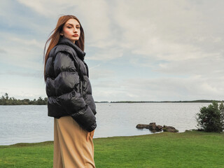 A woman stands on a grassy field and lake with small island in the background. The model is wearing...