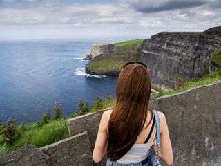 Young girl looking at stunning cliff of Moher, county Clare, Ireland. Travel and tourism. Warm sunny day with cloudy sky. Popular Irish landmark.