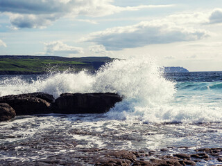 Fototapeta premium Ragged and tough stone coast hit by power full ocean waves, cliff in the background and cloudy sky. Doolin area, county Clare. Ireland. Popular tourist sightseeing area. Irish nature landscape.