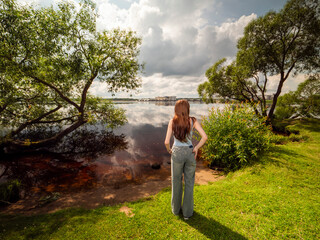 A woman stands by a river, looking out at the trees. The scene is peaceful and serene. Kegums park area, Latvia. Warm sunny day. Model is slim in casual cloths