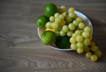 A fresh assortment of fruits, including a bunch of green grapes, bright limes and lemon, sit in a white bowl on a light wooden table. The arrangement is bright and natural.