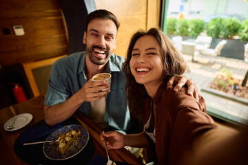 Cheerful couple taking selfie while eating cake and drinking coffee in cafe.