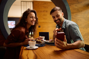Happy couple taking selfie while eating cake in cafeteria.