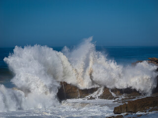 Huge ocean wave crashes against coastal cliff, sending enormous amount of white water and foam high into the air. Water cascades down the face of a scenic cliff and spills over jagged rocks below.