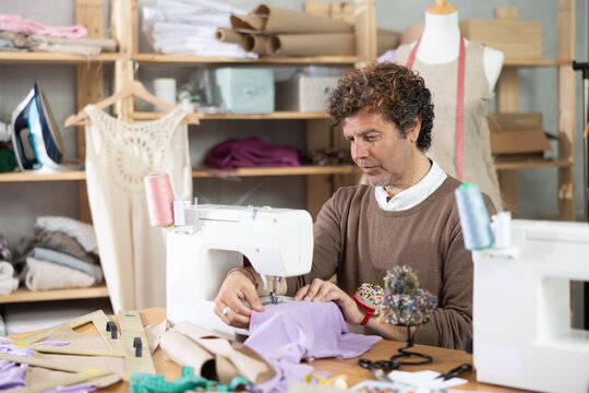 Adult man sews clothes on a sewing machine against the background of shelves with fabrics and a mannequin. Process of sewing a collection of clothes in the workshop