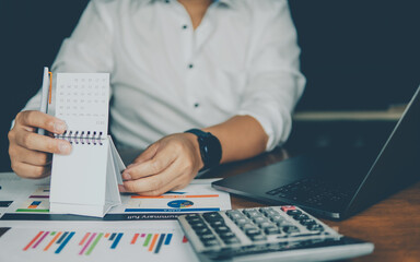 A professional writes on a notepad surrounded by financial charts, a calculator, and a laptop in an office setting.
