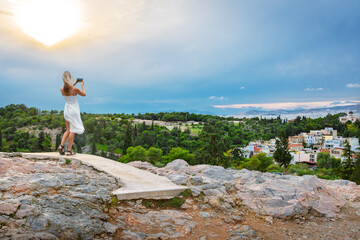 A young woman takes a photo from the top of Areopagus Hill, with the Holy Church of Agia Marina Thisiou and Athens observatory in view, Athens Greece.