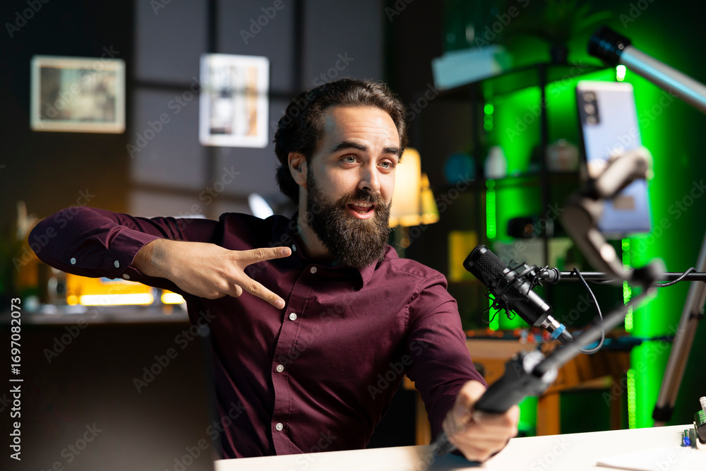 Poster influencer doing peace sign in front on cellphone attached to selfie stick in studio, chatting with 