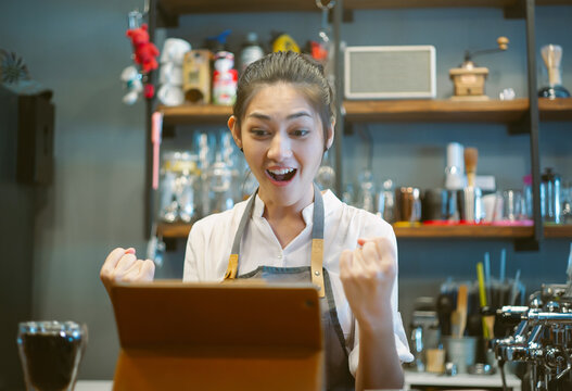 Asian young barista in apron smiles with excitement while celebrating a joyful moment at coffee shop counter, using a tablet and showing happiness in her workplace. Small businesses owner concept.