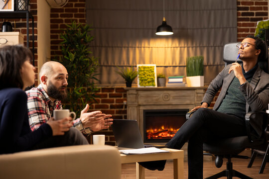 Therapist with pen to chin meets with clients in private clinic, offering guidance for relationship support. African American psychologist observes young couple sharing feelings during session.