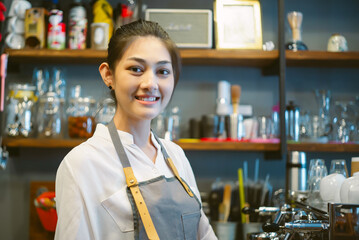 Asian young female barista wearing a white shirt and gray apron smiles warmly behind the counter in...