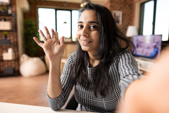 Happy female freelancer sitting in cozy living room waves at phone during video chat. Indian businesswoman seated at home office desk, greeting caller with a smile and friendly hand gesture.