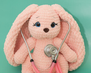 Pink plush knitted bunny listening to heart with pink stethoscope on green background, playing...