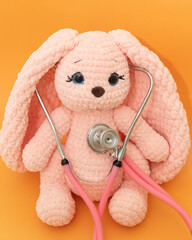 Pink plush knitted bunny listening to heart with pink stethoscope on orange background, playing...