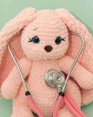 Pink plush knitted bunny listening to heart with pink stethoscope on green background, playing...
