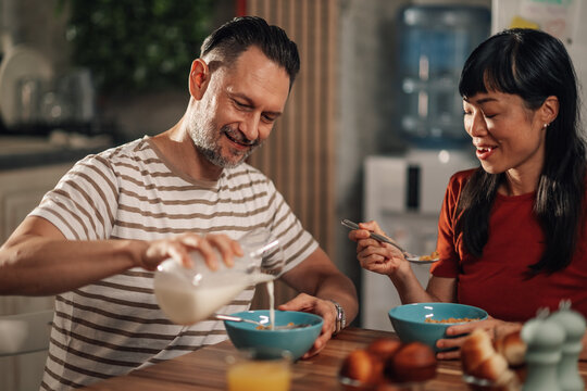 Happy couple enjoying breakfast together in kitchen, pouring milk into cereal bowl