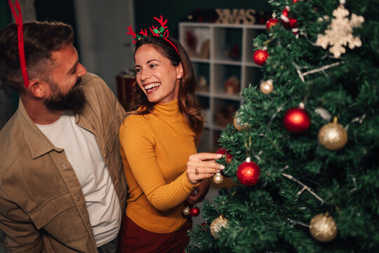 Happy couple decorating christmas tree wearing reindeer antlers headband - Powered by Adobe