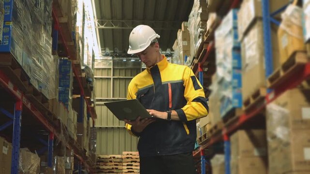 Professional male worker wearing  safety helmet working industrial warehouse storing and distributing logistics goods warehouse center using laptop record and monitor goods on the shelves.