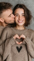 Smiling woman with heart hands, man kisses cheek against neutral background
