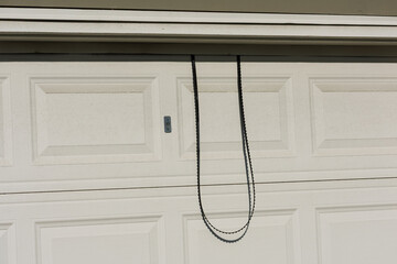 Residential garage door with textured white panels and long black drive belt hanging down in front, illustrating damaged opener hardware and repair requirements.