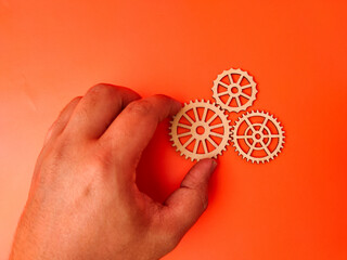 The image shows a hand holding a wooden gear, with two other wooden gears lying on an orange...