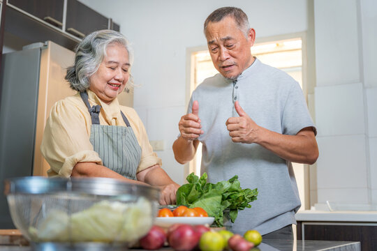 Asian senior couple cooking together in the kitchen, Happy retired couple preparing healthy food, Grandfather gives a thumbs up while grandmother smiles and prepares vegetables - Powered by Adobe