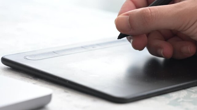Hand uses a stylus pen on a graphic tablet to draw design or retouch images on a computer in a digital creative workspace.