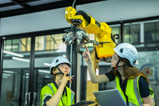 Two female engineers in safety gear collaborate on an industrial robot. They are programming the automated arm with a laptop and controller in a modern smart factory or production plant. - Powered by Adobe