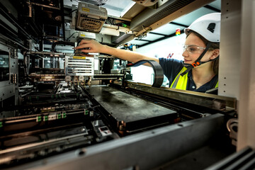 A young female worker in a hard hat carefully works on a large machine in a factory. She is focused on connecting the electronic wires to make the industrial equipment work.