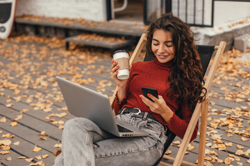 Beautiful woman in cozy outfit works at laptop while sitting on chair near cafe in autumn park. Female student studies information using computer outdoor. Freelancer works remotely outside home.